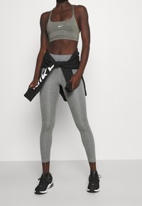 Sportovní outfit zahrnující šedou sportovní podprsenku s bílým logem, ladící šedé legíny a černé tenisky s bílými akcenty. Černý top uvázaný v pase.