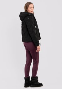 Schwarze Puffjacke mit gestepptem Design, angenommener Kapuze und silbernen Reißverschlüssen. Kombiniert mit burgunderfarbenen Leggings und schwarzen Stiefeln mit umgeschlagenem Saum.