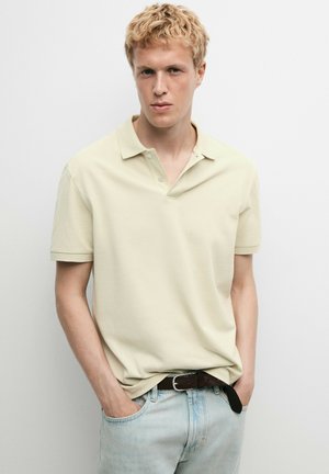 Jeune homme aux cheveux blonds courts portant un polo beige clair et un jean bleu clair, les mains dans les poches, debout devant un fond blanc uni.