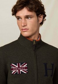 Tmavě zelená zipová mikina s žebrovaným límcem. Obsahuje nášivku Union Jack a logo "HACKETT". Materiál je jemně pletený.