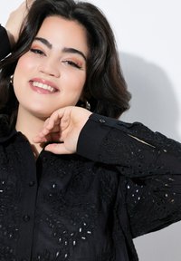 Chemise noire brodée avec des motifs floraux en dentelle, comportant des boutons, des manches longues et un tissu texturé. Finition lisse avec un design ajusté.