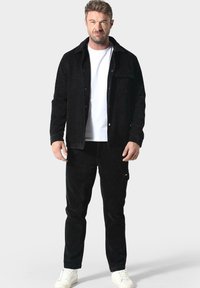 Muž stojící v černé bundě a kalhotách s bílým tričkem a bílými teniskami na jednobarevném světlém pozadí.