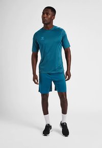 Tealfärgad träningsskjorta och shorts av lätt tyg. Skjortan har rund halsringning och korta ärmar, med logodetalj.