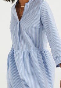 Sininen ja valkoinen raidallinen mekko, jossa on henley-kaula-aukko, pitkät hihat ja kootut vyötärö. Raikas puuvillakangas; polven pituinen hameosa.