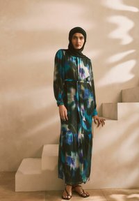 Robe maxi en tissu fluide, avec des motifs abstraits bleus et verts, manches longues, taille cintrée avec un lien, et ourlet en étages. Hijab noir porté.
