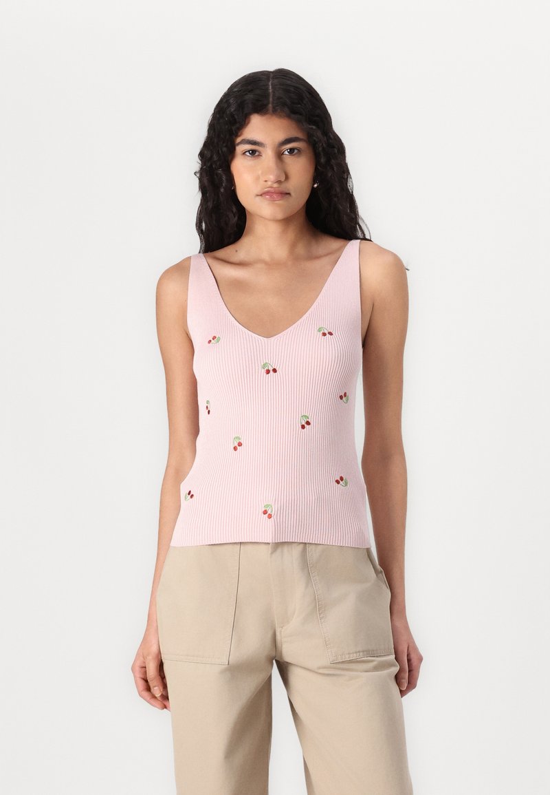 Top a righe rosa con scollatura a V e piccola ricamatura di ciliegia rossa. Indossato con pantaloni beige. Tessuto morbido e design aderente.