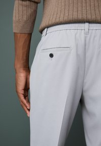 Light gray trousers with a smooth texture, featuring a back pocket with a black button and an elastic waistband. Close-up view shows the side.