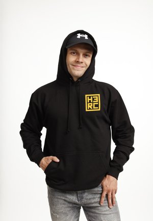 Uśmiechnięty mężczyzna w czarnej bluzie z kapturem z żółtym logo "HERC", czarnej czapce Under Armour i szarych dżinsach, ręce w kieszeniach i po bokach.