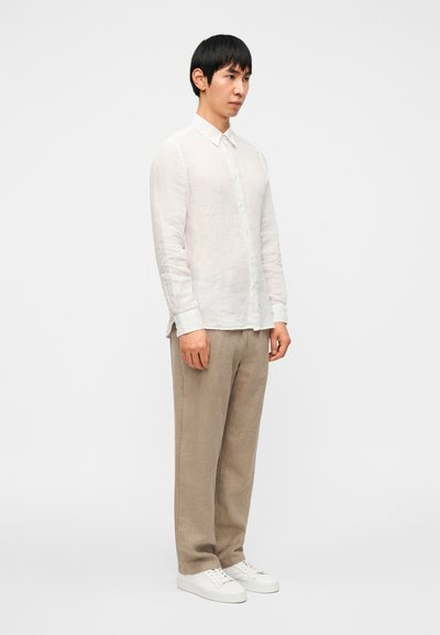 Άσπρο λινό πουκάμισο με κουμπιά, μπεζ λινό παντελόνι και άσπρα αθλητικά παπούτσια. Το μοντέλο στέκεται προφίλ μπροστά σε απλό φόντο.