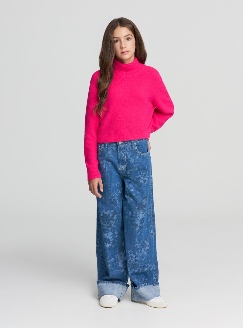 Ragazza con lunghi capelli castani che indossa un maglione rosa brillante, jeans azzurri a gamba larga con motivo floreale e sneakers bianche, in piedi con una mano dietro la schiena.