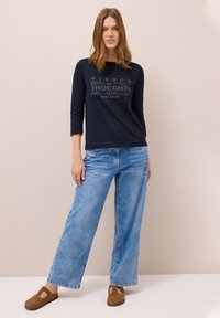 Marineblaues Langarmshirt mit Textdesign, kombiniert mit hellblauen weiten Jeans und braunen Wildleder-Slippern. Der Hintergrund ist neutral.