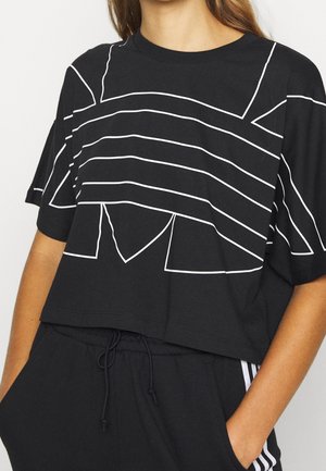 Kobieta nosząca czarny krótki t-shirt z białym abstrakcyjnym wzorem linii oraz czarne spodnie z troczkiem i białymi paskami, ręce w kieszeniach.