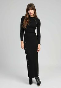 Robe pull côtelée noire avec un col montant, des manches longues et des accents de boutons dorés sur le côté. Fente latérale et silhouette ajustée.