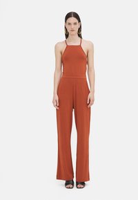 Une combinaison couleur rouille avec un corsage ajusté, des bretelles fines et des pantalons à jambes larges. Tissu doux et lisse avec un design minimaliste.