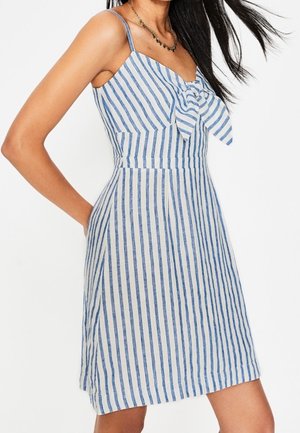 Woman wearing a sleeveless, blue and white vertical striped dress with a front bow detail and thin shoulder straps.
