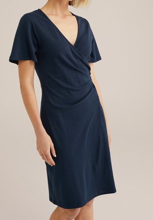 Kobieta ubrana w granatową sukienkę kopertową do kolan z krótkimi falbanianymi rękawami i dekoltem w kształcie litery V, stojąca na beżowym tle.