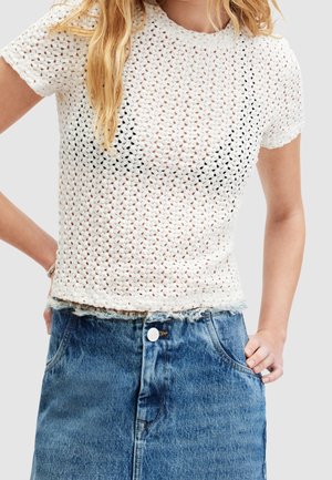 Weißes Strickoberteil mit Mesh-Design, rundem Ausschnitt und kurzen Ärmeln, kombiniert mit einem Jeansrock mit ausgefranstem Saum und vorderen Knöpfen.