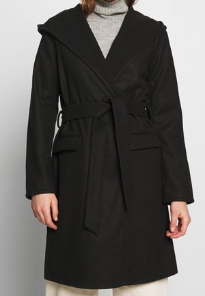 Personne portant un manteau noir ceinturé avec un grand col et des poches à rabat par-dessus un pull à col roulé gris clair.