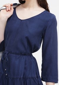 Abito blu navy a maniche lunghe con scollatura a V, vita accentuata e gonna a strati. Presenta un laccio con coulisse e bottoni in vita.