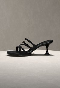 Μαύρο σανδάλι mule με λουράκια, τετράγωνη μύτη και καμπυλωτό χαμηλό τακούνι σε ανοιχτή επιφάνεια σε ουδέτερο φόντο.