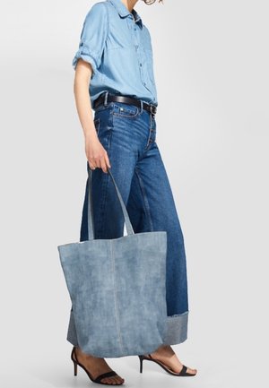 Sac fourre-tout en denim avec tissu bleu clair, design simple, anses robustes et texture lisse. Porté avec une chemise bleu clair et un jean large.