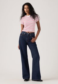 Naine, kellel on pikad lokkis juuksed, kannab valget ja punast triibulist lühikeste varrukatega toppi ning tumesiniseid kõrge vöökohaga laiade säärtega teksapükse.