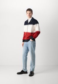 Giacca con zip in blu navy, bianco e rosso, con strisce orizzontali, polsini elasticizzati e un'etichetta. Abbinata a jeans azzurri e scarpe nere.