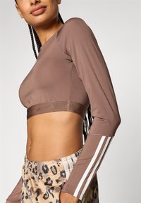 Brun langermet crop-top laget av glatt stoff, med en teksturert midjebånd og hvite akcentstriper langs ermene. Leopardmønstrede bukser.