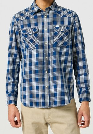 Hombre con camisa de manga larga a cuadros azul y blanca con dos bolsillos en el pecho y pantalones beige, de pie contra un fondo liso.