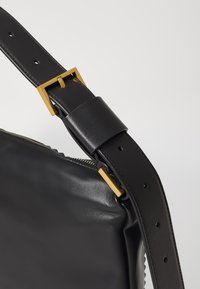 Cinghia nera in pelle con hardware dorato, caratterizzata da una texture liscia, una fibbia regolabile e dettagli rivettati. Cerniera visibile sul lato della borsa.