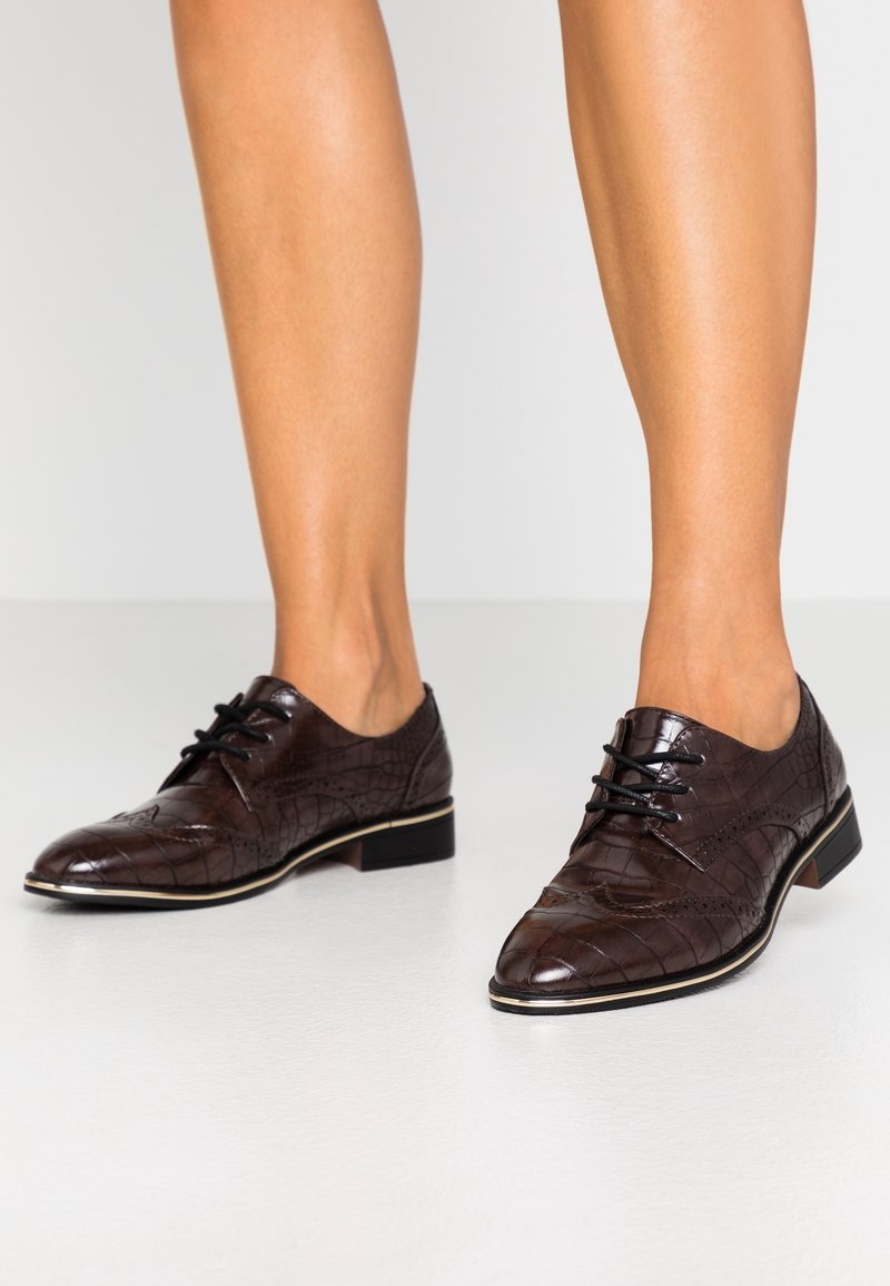 Chaussures Oxford en cuir marron à motif crocodile, à lacets, portées sur des jambes nues sur un fond clair.