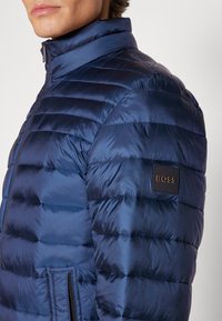 Chaqueta acolchada azul marino con textura acolchada, cuello alzado, cierre de cremallera y un parche con el logotipo en el brazo izquierdo.