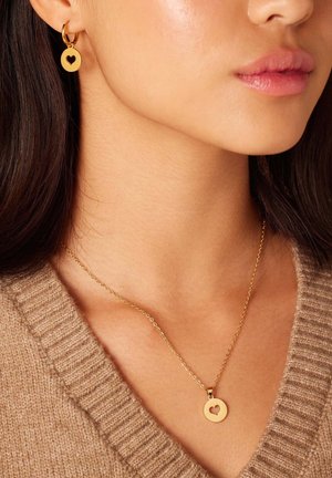 De gouden sieradenset bestaat uit oorringen en een ketting met cirkelvormige hangers met hartvormige uitsparingen, gedragen over een lichtbruin gebreid vest.
