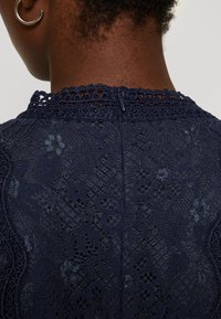 Marinblå spetsklänning med blommönster, texturerad livstycke och hög krage med scalloped kant. Bakre dragkedja synlig.