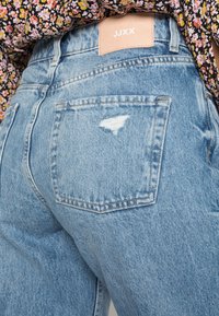 Jeans de denim azul claro com um bolso desgastado e uma etiqueta de couro bege com a inscrição "JJXX." Apresenta um corte reto e detalhes de costura visíveis.