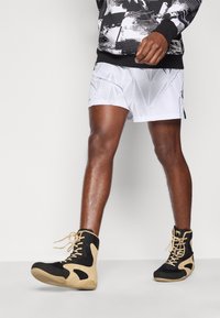 Μαύρα και μπεζ sneakers υψηλού τύπου με διαπνέον ζωντανό πλέγμα και δερμάτινες λεπτομέρειες, σε συνδυασμό με λευκά σορτς που έχουν γεωμετρικό μοτίβο φύλλων.