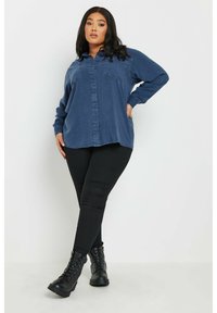 Camicia di jeans di un blu scuro con chiusura a bottoni e due tasche sul petto, abbinata a leggings neri e stivali neri con lacci alla caviglia.