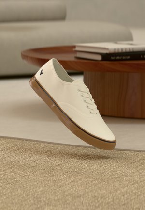Hvid lærredssneaker med brun gummisål og sort logo flydende over beige tæppe, med træ sofabord og bøger i baggrunden.