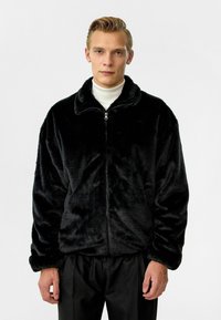 Zwarte faux fur zip-up jas met een zachte, gestructureerde oppervlakte; heeft een hoge kraag en een relaxte pasvorm. Draag het over een witte coltrui.