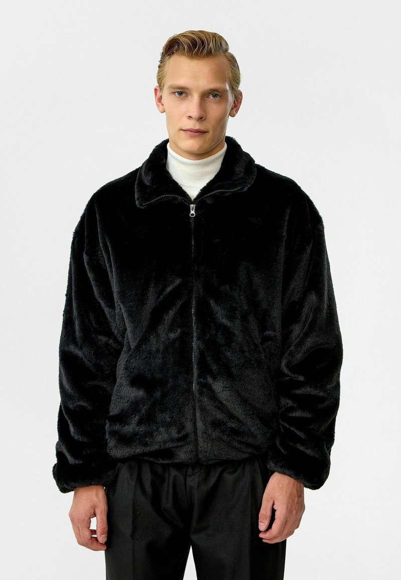Zwarte faux fur zip-up jas met een zachte, gestructureerde oppervlakte; heeft een hoge kraag en een relaxte pasvorm. Draag het over een witte coltrui.