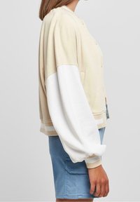 Beige bomberová bunda s bílými objemnými rukávy, žebrovanými manžetami a zapínáním na knoflíky. Tkanina vypadá měkce a má texturu, doplněná o světle modré kraťasy.