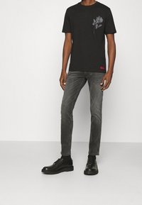 T-shirt nera a maniche corte con una grafica floreale grigia sul lato sinistro del petto, abbinata a jeans grigi sbiaditi e scarpe nere con lacci.