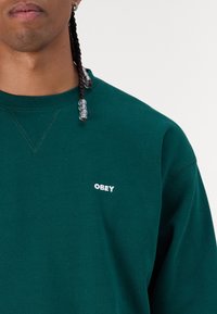 Green sweatshirt with a crew neck, featuring a small white "OBEY" logo on the left chest and a textured fabric finish.