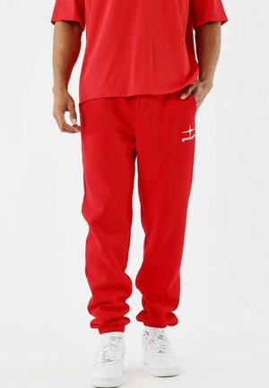 Hombre vestido con pantalones de chándal rojos y una camiseta roja a juego, con zapatillas blancas, de pie contra un fondo blanco plano.