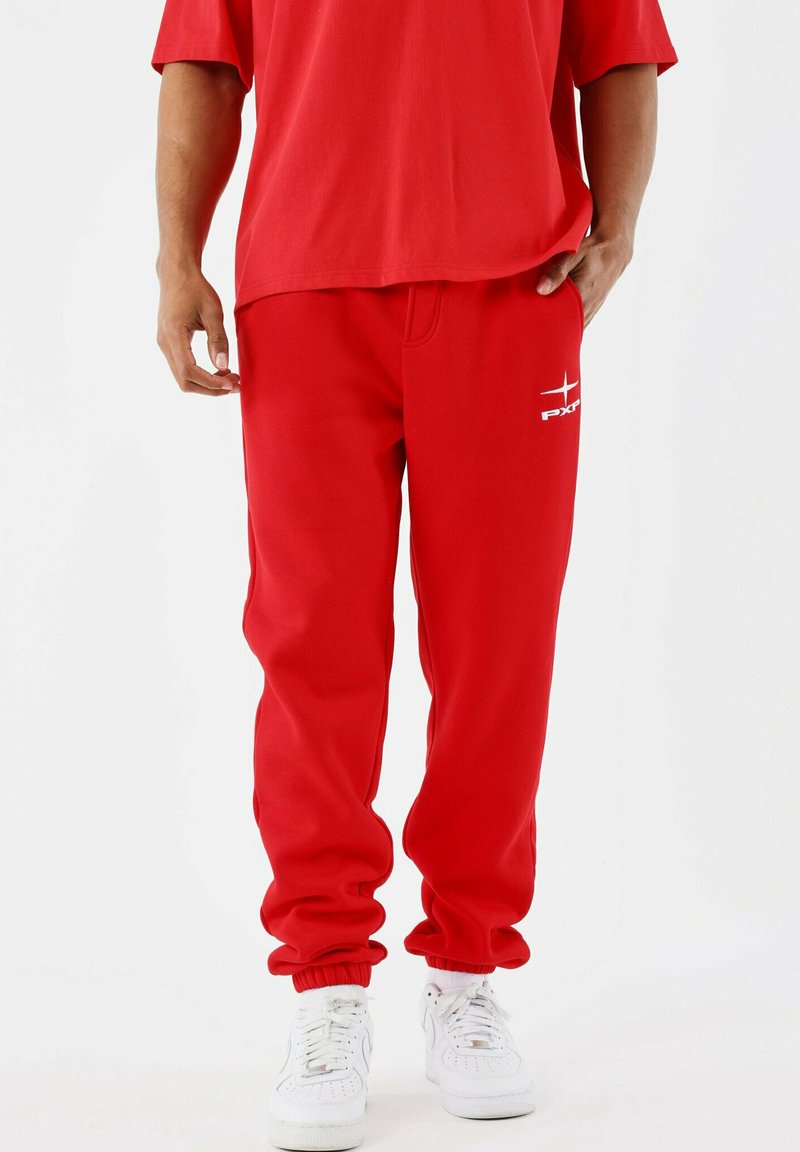 Man in rode sweatpants en een bijpassend rood shirt met witte sneakers, staand tegen een effen witte achtergrond.