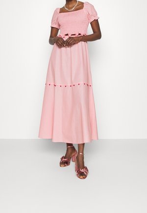 Kobieta w różowej, midi sukience z bufkami na rękawach, z marszczonym gorsetem i ozdobą z pomponów, w połączeniu z błyszczącymi różowymi szpilkami ozdobionymi kokardkami.