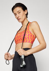 Träningsbh i rosa med gult abstrakt mönster, med en rundad halsringning. Modellen håller ett svart hopprep med grå handtag. Svarta högmidjade leggings.