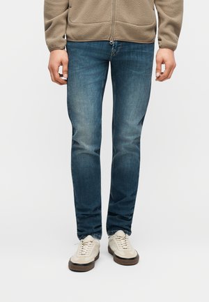 Άνδρας που φορά ξεθωριασμένο μπλε τζιν, μπεζ φλις μπουφάν και λευκά αθλητικά παπούτσια, στέκεται μπροστά σε λευκό φόντο.