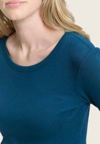 Detailní záběr ženy v tyrkysovém topu s dlouhými rukávy, který ukazuje výstřih, klíční kost a část jejích blond vlasů.