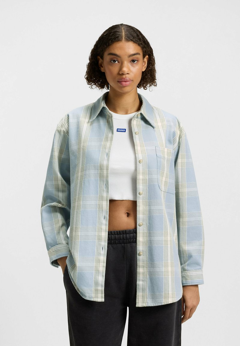 Lyseblå ternet flannel skjorte med grønne detaljer, knapstolpe foran, enkelt brystlomme, båret over en hvid cropped top og sorte bukser.